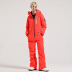 Model wearing the orange red Alpine Access one-piece ski suit, with hood down and hands in the pockets.